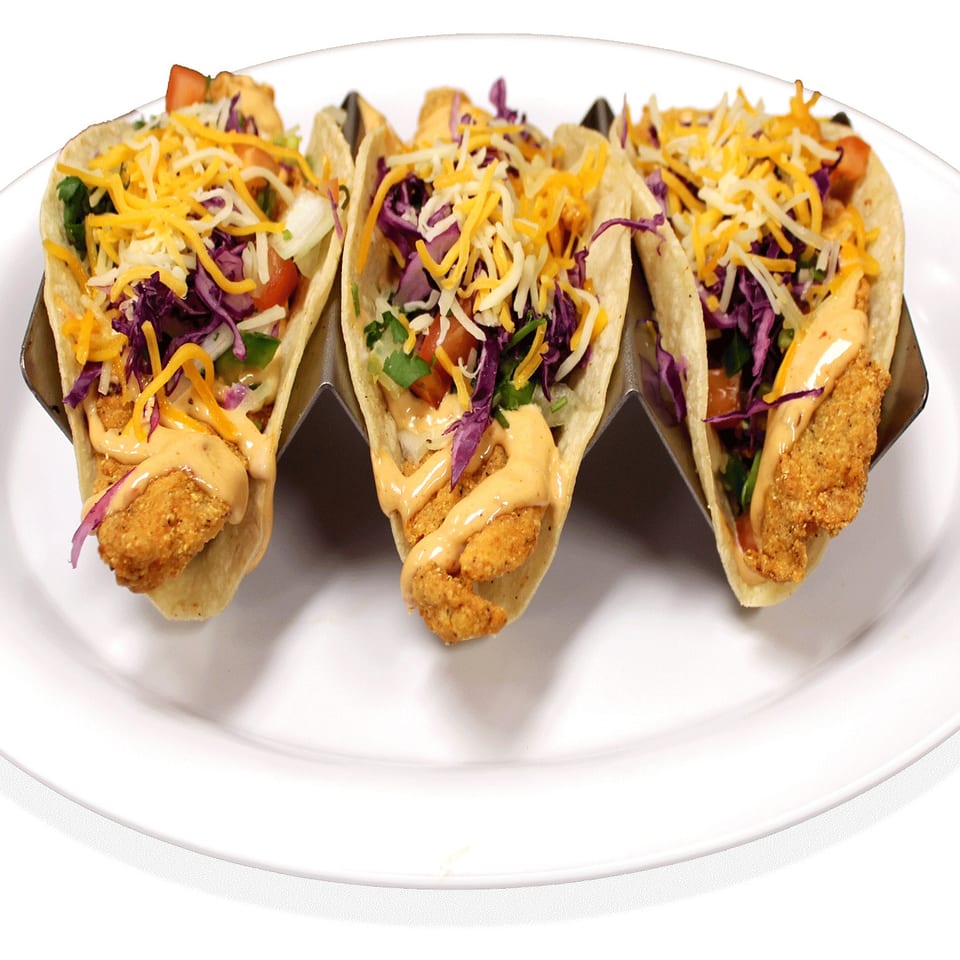 Three tacos with crispy fried protein, shredded cheese, red cabbage, and creamy sauce on a white plate.